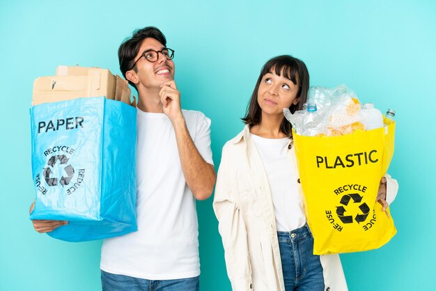 Jovem casal segurando um saco cheio de plástico e papel para reciclar isolado em fundo azul pensando em uma ideia enquanto coça a cabeça