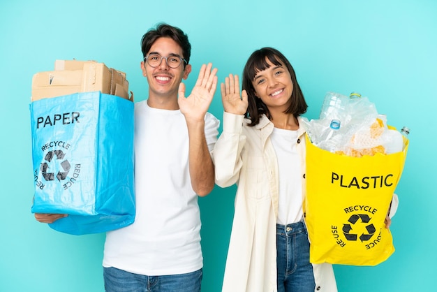 Jovem casal segurando um saco cheio de plástico e papel para reciclar isolado em fundo azul saudando com a mão com expressão feliz