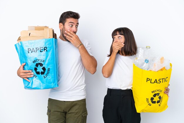 Jovem casal segurando um saco cheio de plástico e papel para reciclar isolado no fundo branco, cobrindo a boca com as mãos para dizer algo inapropriado