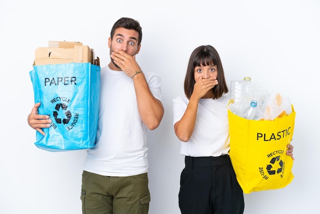 Jovem casal segurando um saco cheio de plástico e papel para reciclar isolado no fundo branco, cobrindo a boca com as mãos para dizer algo inapropriado