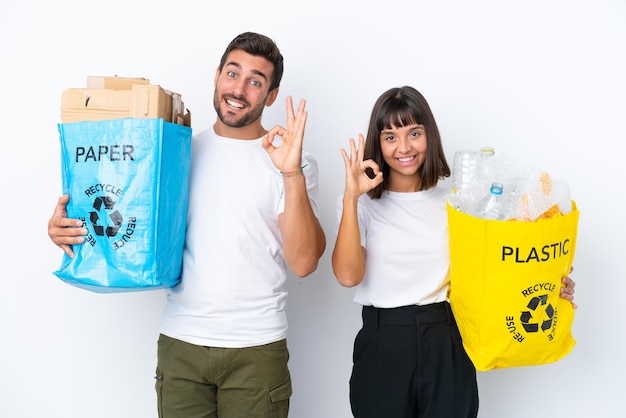 Jovem casal segurando um saco cheio de plástico e papel para reciclar isolado no fundo branco, mostrando um sinal de ok com os dedos