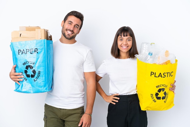 Jovem casal segurando um saco cheio de plástico e papel para reciclar isolado no fundo branco posando com os braços no quadril e sorrindo