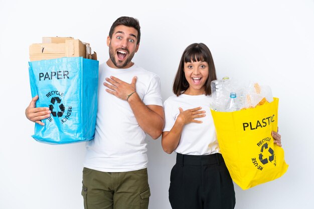 Jovem casal segurando um saco cheio de plástico e papel para reciclar isolado no fundo branco surpreso e chocado ao olhar para a direita