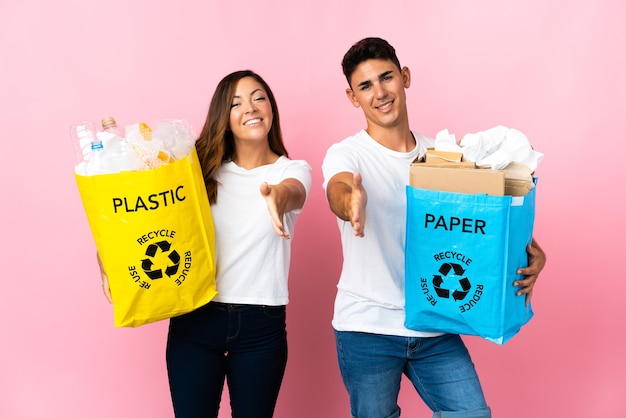 Jovem casal segurando uma sacola cheia de plástico e papel na rosa apertando as mãos para fechar um bom negócio