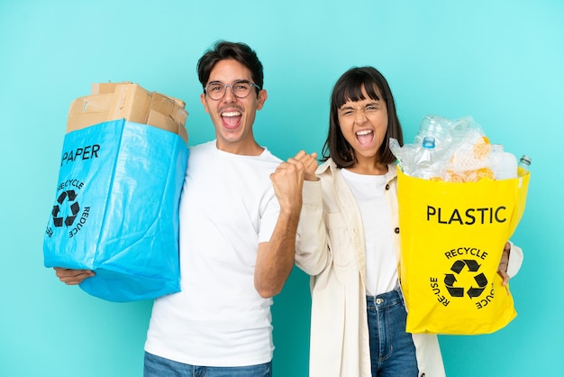 Jovem casal segurando uma sacola cheia de plástico e papel para reciclar isolada em um fundo azul, comemorando uma vitória