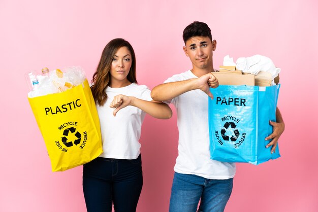 Jovem casal segurando uma sacola cheia de plástico e papel rosa mostrando o polegar para baixo com expressão negativa