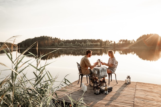 Jovem casal sentado à mesa passa algum tempo juntos no cais de madeira no lago da floresta, comemorando o aniversário, bebendo vinho. O amor está no ar, o conceito de história de amor. Encontro romântico no lago com velas.