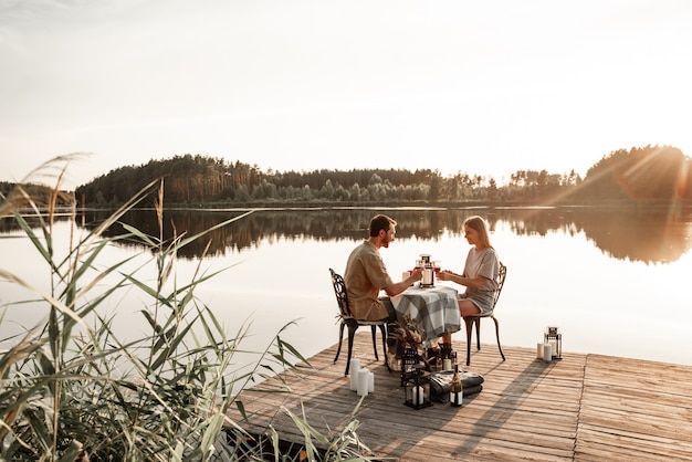 Foto jovem casal sentado à mesa passa algum tempo juntos no cais de madeira no lago da floresta, comemorando o aniversário, bebendo vinho. o amor está no ar, o conceito de história de amor. encontro romântico no lago com velas.