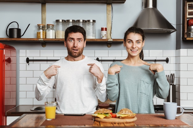 Jovem casal sentado na cozinha durante o café da manhã em casa, apontando o dedo um para o outro