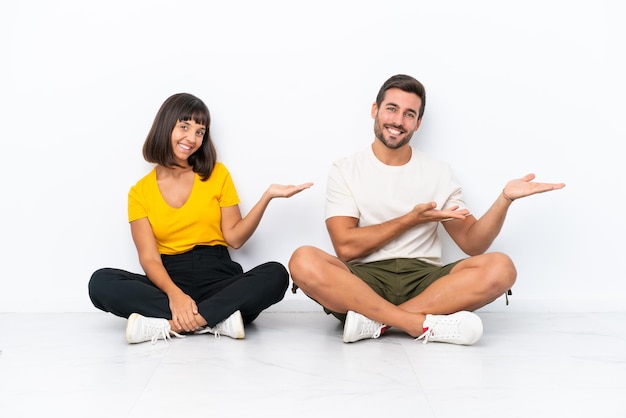 Jovem casal sentado no chão, isolado no fundo branco, apontando para trás e apresentando um produto