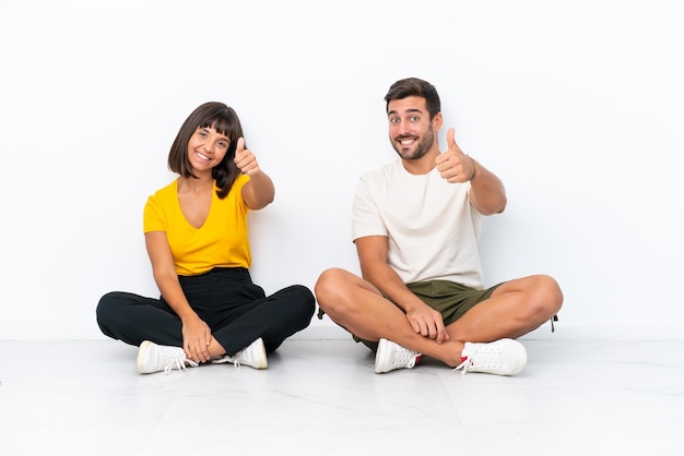 Foto jovem casal sentado no chão isolado no fundo branco dando um polegar para cima gesto porque algo bom aconteceu