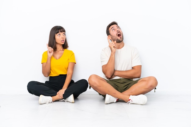 Foto jovem casal sentado no chão isolado no fundo branco pensando em uma ideia apontando o dedo para cima