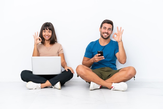 Jovem casal sentado no chão segurando pc e celular isolado no fundo branco, mostrando um sinal de ok com os dedos