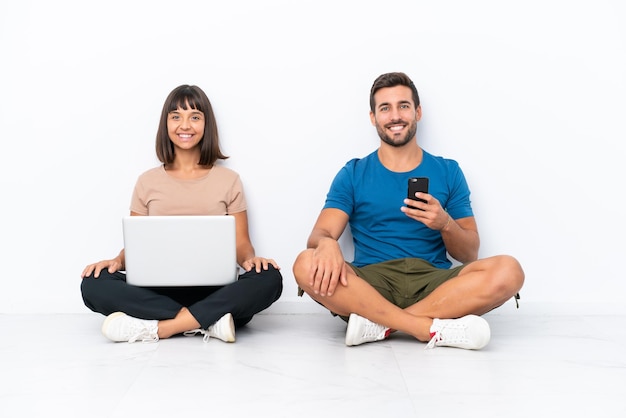 Jovem casal sentado no chão segurando pc e celular isolado no fundo branco posando com braços no quadril e sorrindo