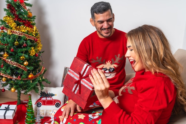 Jovem casal sentado no sofá com decoração e roupas vermelhas de Natal namorado dando um presente para a namorada e namorada muito feliz