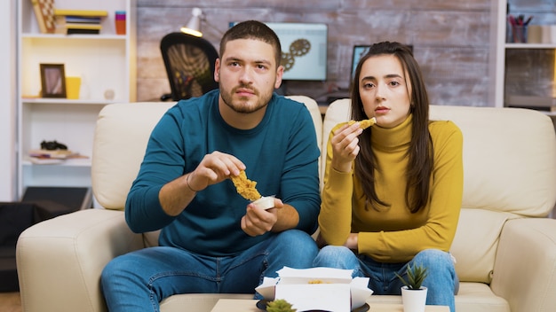 Jovem casal sentado no sofá comendo frango frito enquanto assiste tv.