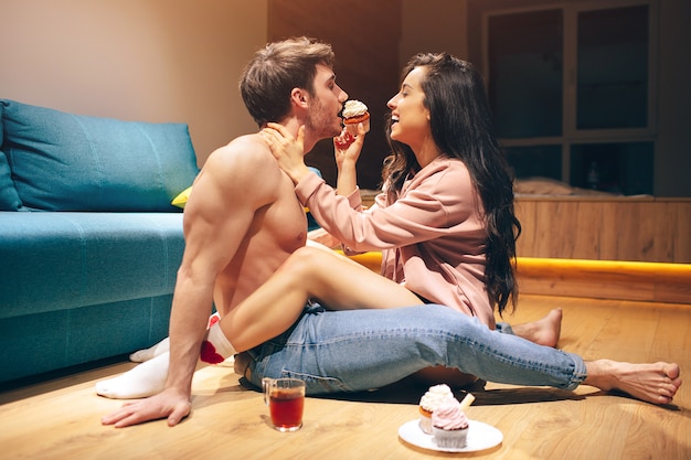 Jovem casal sexy tem intimidade na cozinha à noite. Mulher linda gostosa alimentando o homem com alimentos doces e panquecas. Pernas cruzadas. Panquecas e chá no chão.