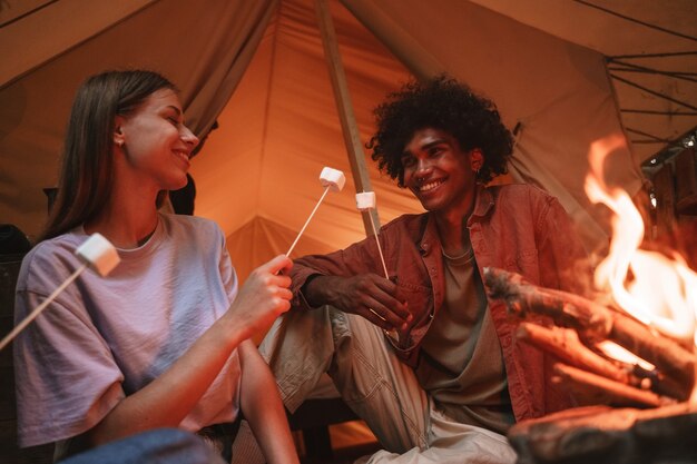 Jovem casal sorridente assando marshmallows no espeto sobre a fogueira no acampamento, olhando um para o outro com amor, desfrutando de glamping ao ar livre. União de férias reaberta após bloqueio pandêmico