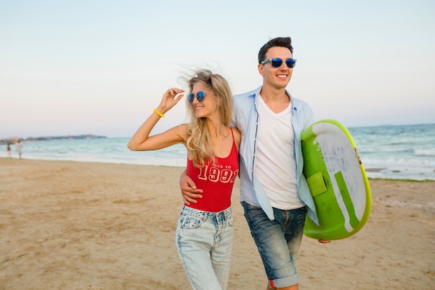 Jovem casal sorridente se divertindo na praia caminhando com prancha de surf