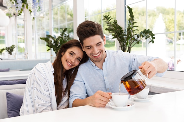 Jovem casal sorridente, sentado à mesa do café, bebendo chá