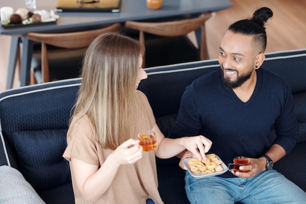 Foto jovem casal sorridente tomando chá com biscoitos e discutindo notícias e fofocas em casa