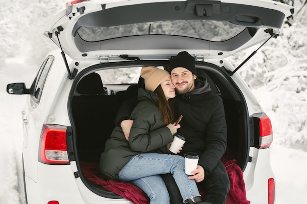 Jovem casal um homem e uma mulher estão sentados no porta-malas de um carro em uma floresta de neve de inverno