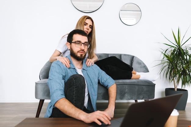 Jovem casal usando laptop, navegando informações e olhando para a tela do laptop na moderna sala de estar em um sofá.