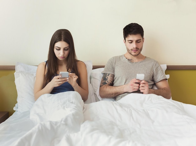 Jovem casal usando o celular, sentado na cama, ignorando um ao outro