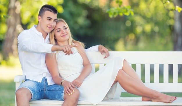 Jovem casal vestido com roupas leves, sentado em um banco branco em um parque, olhando na mesma direção. Menino apontando com o dedo, enquanto o casal está sorrindo.