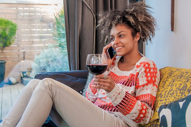 Jovem casual usando o telefone móvel enquanto está sentado no sofá em casa e desfruta de um copo de vinho. Mulher afro-americana falando em um telefone celular