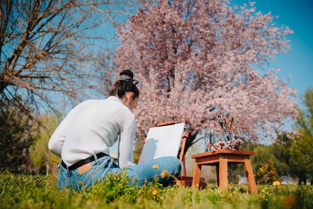 Jovem caucasiana com cabelo ondulado pintando uma tela com tintas rosa pastel em pé na natureza de um parque