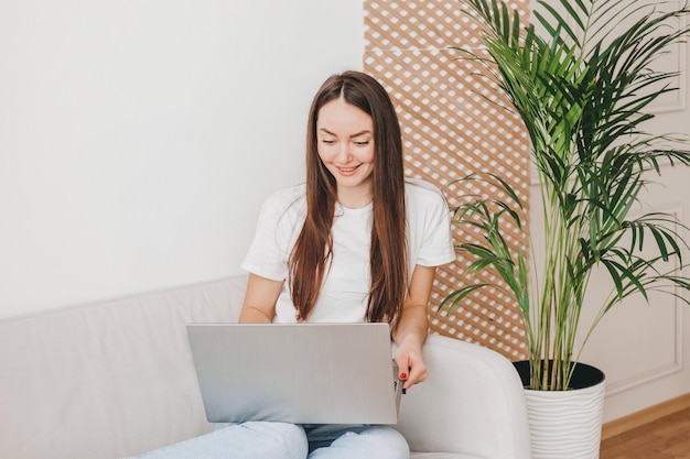 Jovem caucasiana olhando para a tela do laptop sentada no sofá trabalhando e sorrindo