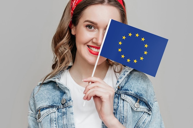 Jovem caucasiana segurando pequena bandeira com emblema da União Europeia isolada em fundo cinza