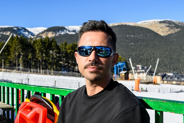 Jovem caucasiano com óculos escuros, piercings e suéter preto, sentado tomando sol na neve.