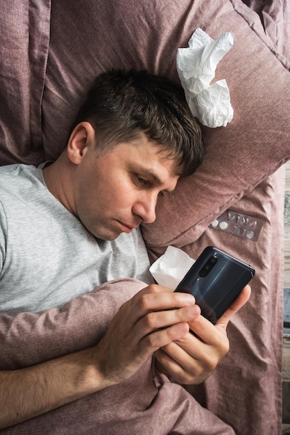 Jovem caucasiano doente com gripe ou vírus usando telefone celular ou smartphone deitado na cama em casa