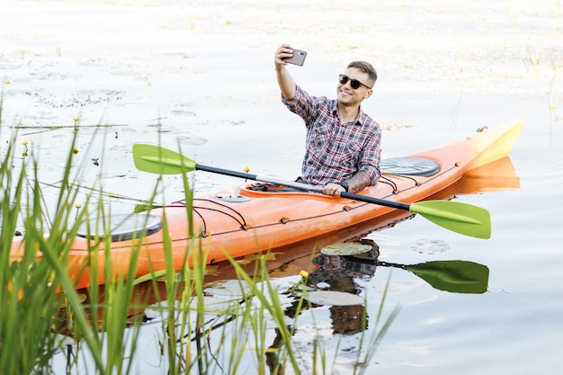 Jovem caucasiano sentado em um caiaque e tirando uma selfie em um telefone celular O conceito de entretenimento aquático
