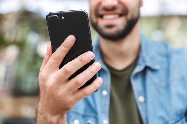 Jovem caucasiano usando fones de ouvido segurando e falando no smartphone enquanto está sentado em um café ao ar livre na cidade