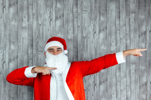 Jovem caucasiano vestido de Papai Noel, apontando com as duas mãos para a direita. Copie o espaço acima