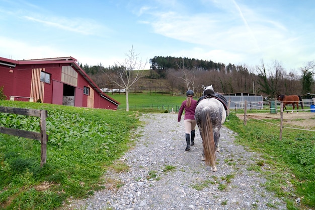 Jovem cavaleiro caminha com seu cavalo branco em um caminho de cascalho com um celeiro vermelho rústico e campos verdes