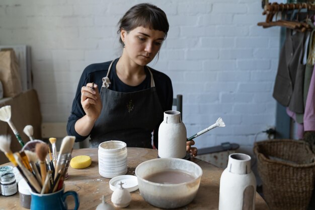 Jovem ceramista concentrada senta-se à mesa na oficina de cerâmica criando jarros e vasos
