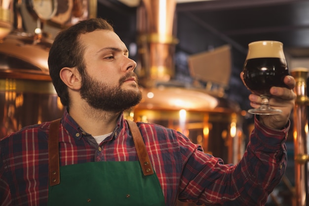 Jovem cervejeiro barbudo vestindo avental trabalhando em sua cervejaria, examinando a cerveja escura em um copo