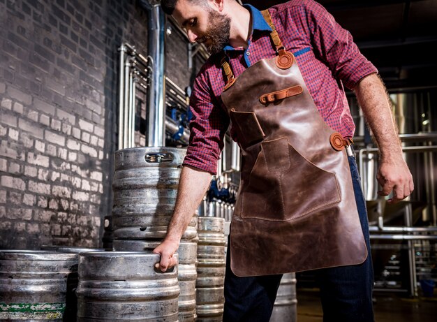 Jovem cervejeiro com avental de couro segurando um barril com cerveja artesanal em uma moderna fábrica de cerveja
