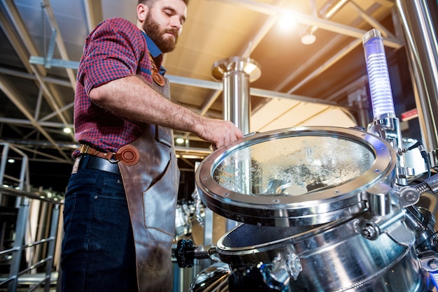 Jovem cervejeiro com avental de couro supervisionando o processo de fermentação da cerveja em uma cervejaria moderna.