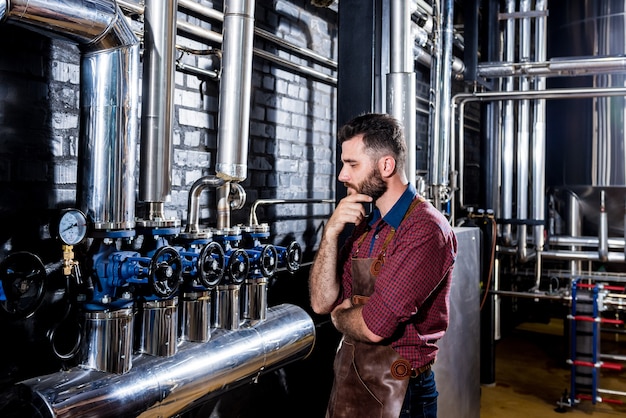 Jovem cervejeiro com avental de couro supervisionando o processo de fermentação da cerveja em uma fábrica de cerveja moderna.