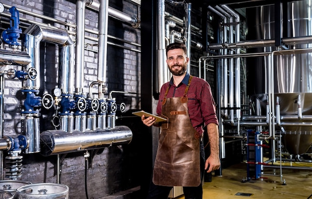 Jovem cervejeiro com avental de couro supervisionando o processo de fermentação da cerveja em uma moderna fábrica de ...