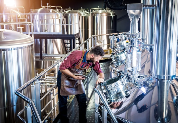 Jovem cervejeiro com avental de couro supervisionando o processo de fermentação da cerveja em uma moderna fábrica de ...