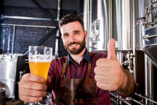 Jovem cervejeiro vestindo um avental de couro está testando cerveja em uma cervejaria moderna