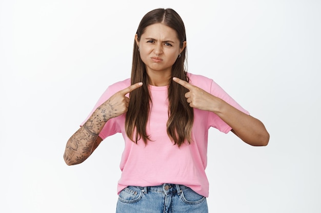 Jovem cética apontando para si mesma e fazendo careta descontente, não gosta de gesto, de pé na camiseta rosa contra fundo branco