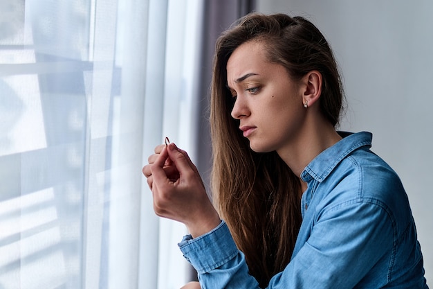 Jovem chateada deprimida solitária mulher divorciada com olhos tristes em uma camisa mantém o anel de ouro e senta-se sozinha em casa durante a preocupação com o casamento fracassado após terminar o relacionamento e o divórcio