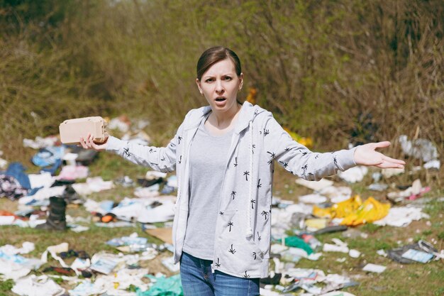 Foto jovem chateada mulher irritada em roupas casuais, limpando segurando o lixo e espalhando as mãos no parque desarrumado. problema de poluição ambiental. pare o lixo da natureza, o conceito de proteção do meio ambiente.